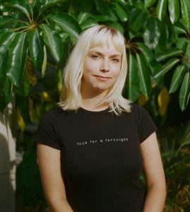A blond white woman in a black tshirt smiles at the camera in front of some green leaves